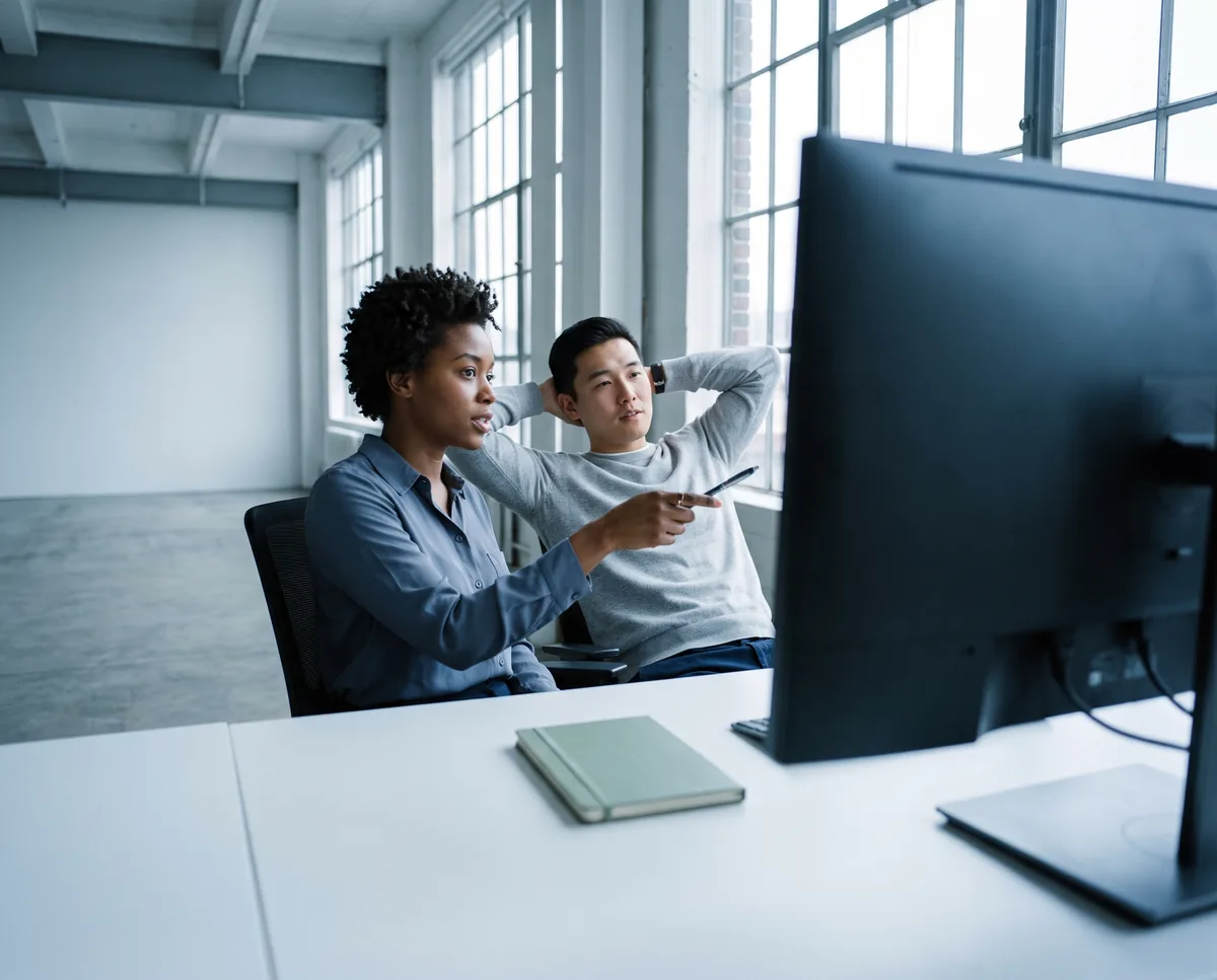 Two teammates at a shared desk, one pointing at a monitor as the other listens